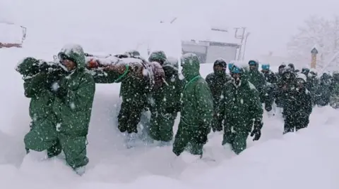 Indian Army via Reuters Several rescuers from the Indian Army, dressed in green overalls, trek through heavy snow after an avalanche struck a camp near Mana village in Uttarakhand state, India
