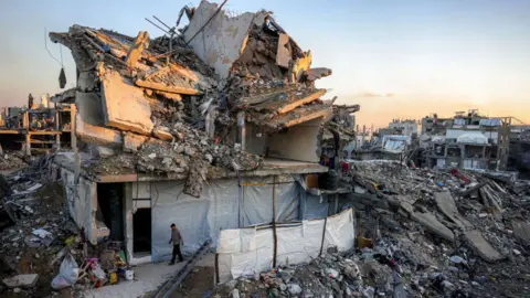 Getty Images The remnants of a collapsed building can be seen with a man walking in front, with the rest of the buildings around them almost all completely flattened