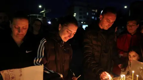 Reuters Four adults and a child stand at a pit lighting candles. Others can be seen standing behind them. The image has been taken at night-time and is dark