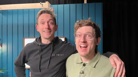 John Robins wearing a black hoodie smiling with his arm around Elis James, wearing a green shirt and glasses, who also smiles. You can see the blue panelled walls of the podcast recording studio behind them