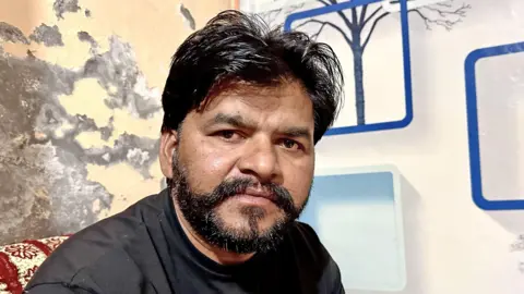 BBC Gurpreet Singh, an Indian man with a beard and black hair that is cut short at the back and sides but long on top, wearing a black T-shirt as he sits in a room with a mural on one wall featuring trees and geometric shapes, while on the other wall the plaster is crumbling.