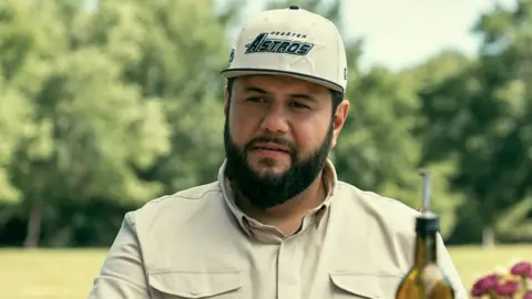 Eddy Chen/Netflix A still from the Netflix show Mo, showing the main character Mo wearing a cream coloured shirt and hat labelled "Houston Astros"as he gazes into the distance with the top of an olive oil bottle in front of him.