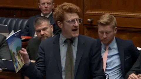 BBC Matthew O'Toole holding the programme for government paper as he speaks to the assembly in Stormont. He has ginger hair, wears clear glasses, and is wearing a dark coloured suit. 