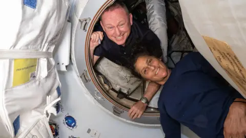 NASA Nasa astronauts Butch Wilmore and Suni Williams onboard the international space station smiling as they pose in the orbiting lab. Butch is looking out from within a white circular hatch, while Suni is lying just outside it. 