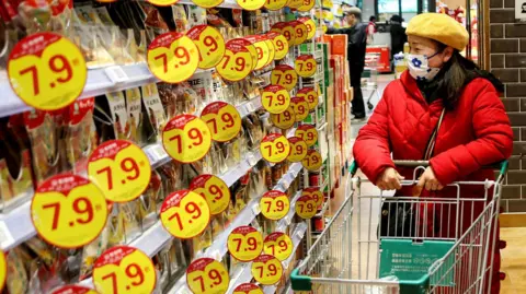 Getty Images A customer wearing a mask and winter jacket browses through products at a supermarket in Lianyungang, in eastern China's Jiangsu province on January 9, 2025. She is standing in an aisle with a trolley in front of her, flanbked by shelves with yellow-colour discount promos on them. 